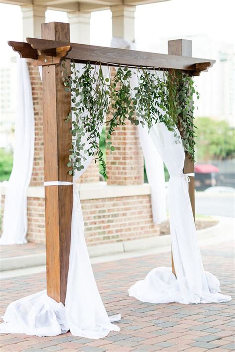 An Outdoor Wedding Ceremony With White Draping And Greenery On The Arbors