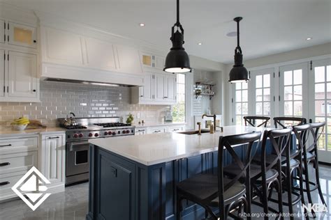 Navy kitchen features a white paneled range hood, flanked by stacked floating rustic wood shelves illuminated by white barn sconces, placed above a stainless steel stove situated next to a wall of navy pantry cabinets enclosing a stainless steel refrigerator. Navy & White Kitchen - NKBA