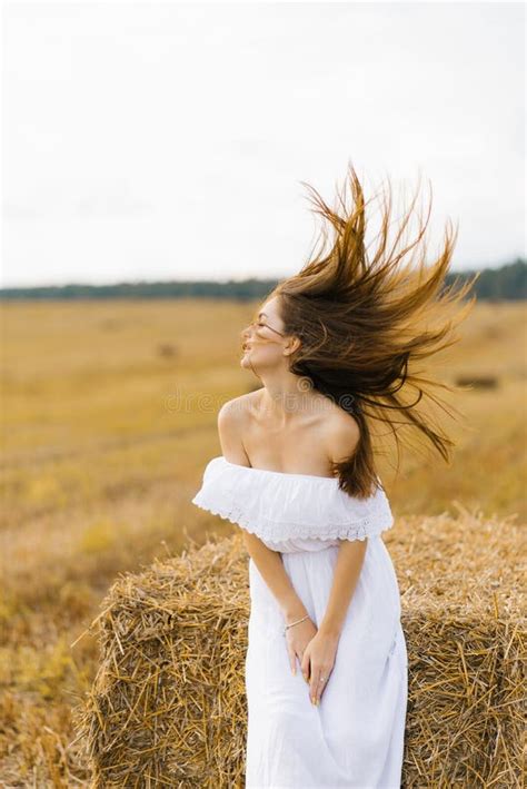 en vacker flicka med blond hår i en vit klänning sitter vid en stråstack på fältet vind i håret