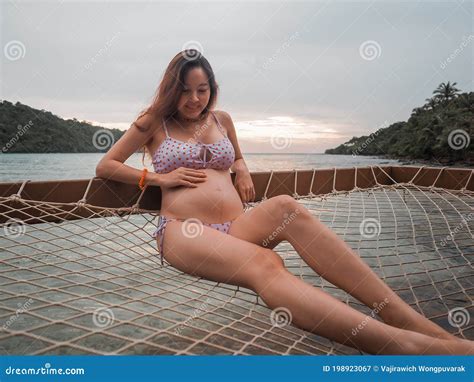 Asian Pregnant Woman In Bikini Sitting On Hammock In Turquoise Sea Water Near Beach Tropical