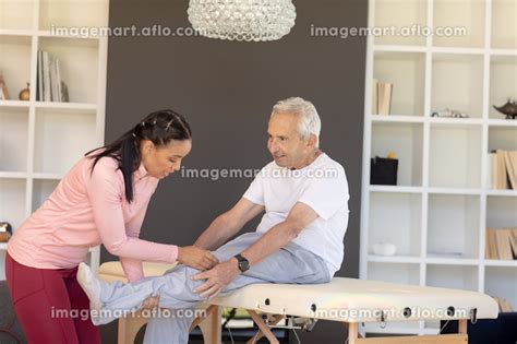 Biracial Female Physiotherapist Giving Leg Massage Therapy To Caucasian Senior Man At Home