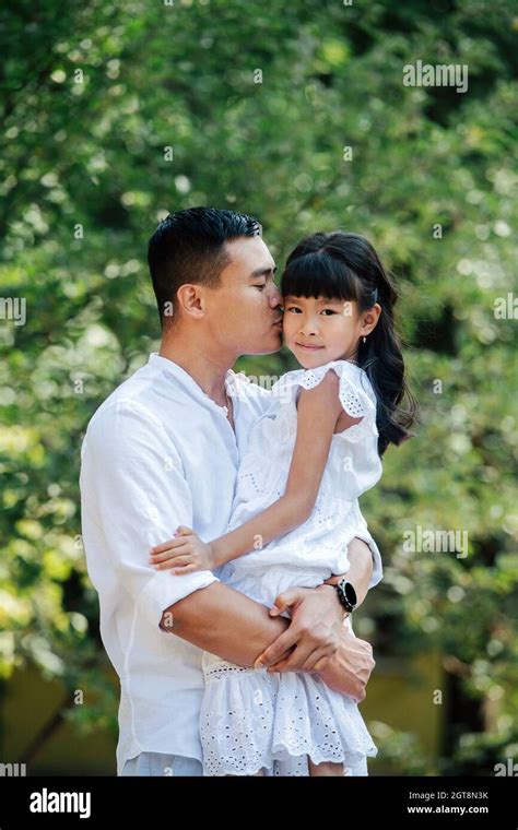 Loving Asian Father Holding His Daughter In His Hands Kissing Her In A Cheek She S Looking At
