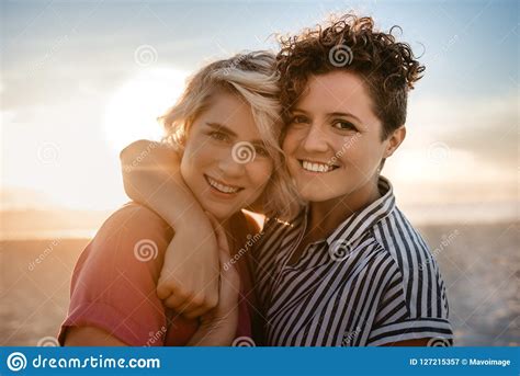smiling lesbian couple standing together on a beach at sunset stock