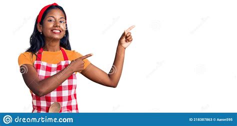 Young Indian Girl Wearing Professional Baker Apron Smiling And Looking At The Camera Pointing