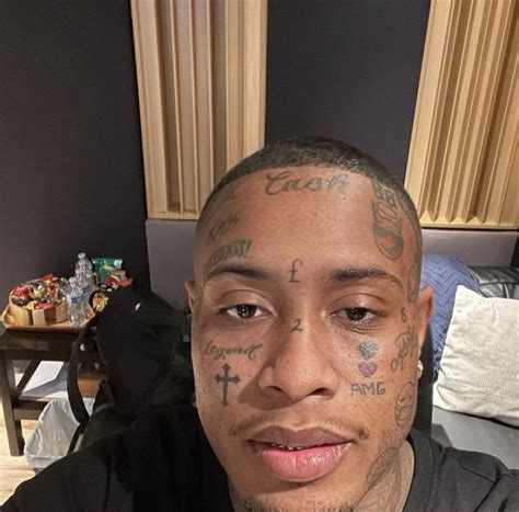 A Man With Tattoos On His Face Sitting In Front Of A Bed And Looking At