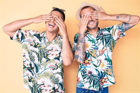 Young Gay Couple Of Two Men Wearing Summer Hat And Hawaiian Shirt Covering Eyes With Hands