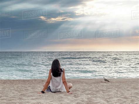 Rear View Of Woman Looking At Sea While Relaxing On Sand Against Cloudy