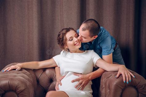 Pregnant Wife Sits In Soft Armchair And Husband Stands Nearby Stock