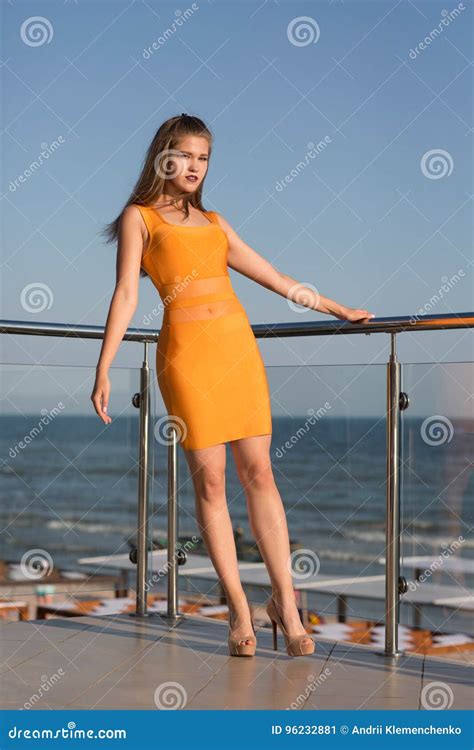A Beautiful Girl Posing On A Balcony On A Blue Sky Background A Lady In A Short Orange Dress