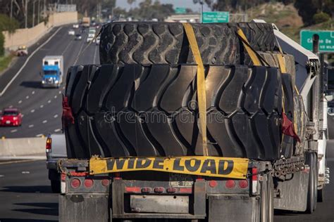 Heavy Equipment Tires Being Hauled On A Flatbed Trailer With Straps And A A Wide Load Sign Stock