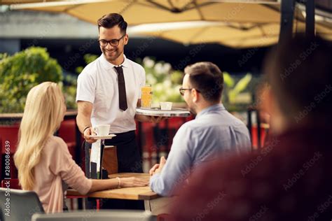 Young Smiling Waiter Serving Coffee To Guests In A Cafe Stock Photo