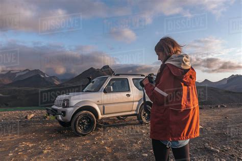 Young Woman Looking Down At Camera In Front Of Car In Mountains Stock