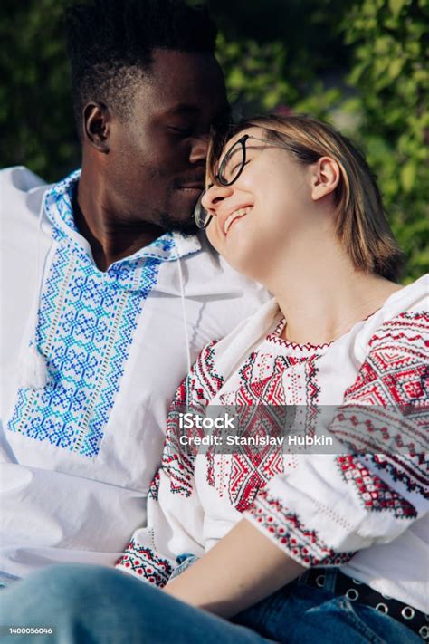 Interracial Happy Couple Sits On Bench In Garden Dressed In Ukrainian Embroidered Shirts Stock