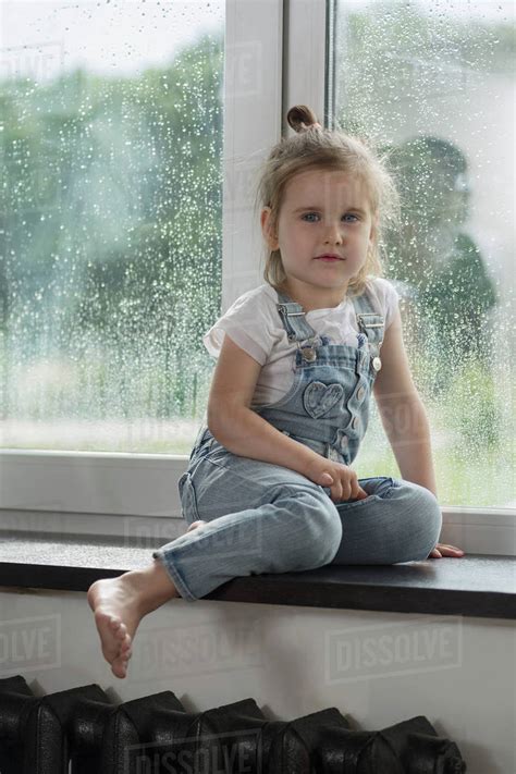Portrait Of Cute Girl Sitting On Window Sill At Home During Rainy