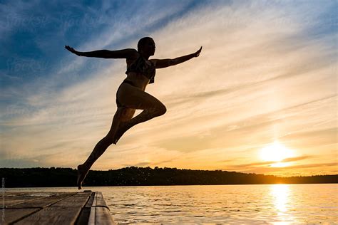 Woman Jumping Off Of Dock Into Warm Summer Cottage Lake At Sunset By