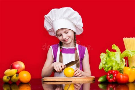 Happy Little Girl In Chef Uniform Cuts Fruit In Kitchen Stock Photo