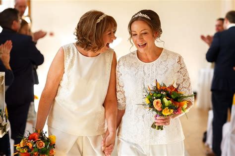 Two Brides Walking Down The Aisle Holding Hands And Smiling At Each Other With Flowers In Their Hand