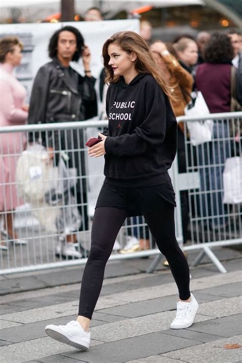 A Woman In Black Sweatshirt And Leggings Walking On Sidewalk With People Behind Her