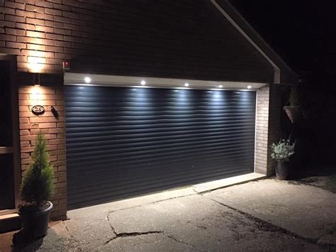 A Garage Door Lit Up At Night With The Lights On And Some Plants In Front