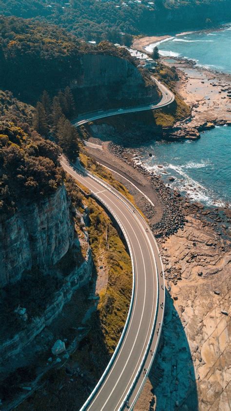 An Aerial View Of A Road Near The Ocean And Cliffs With Trees On Both Sides