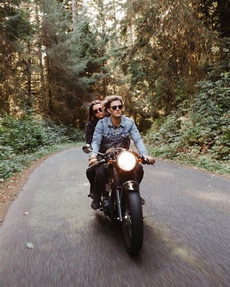 Two People Riding On The Back Of A Motorcycle Down A Road In Front Of Trees