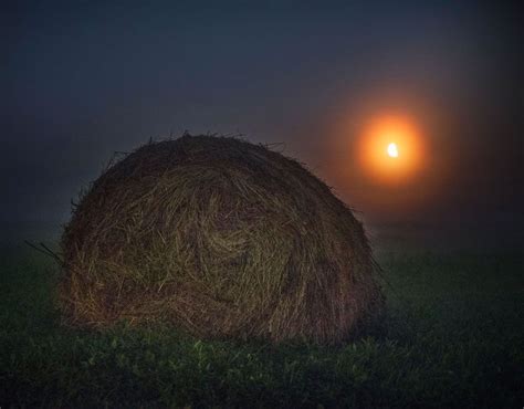 The Moon Lights Up The Night Sky Behind A Barrel Of Hay On A Field