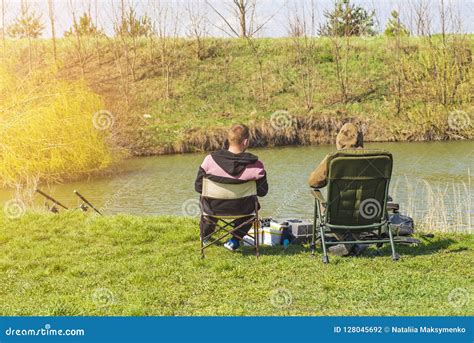 Two Men Sit In Chairs Near The River Fishing With Fishing Rods Stock