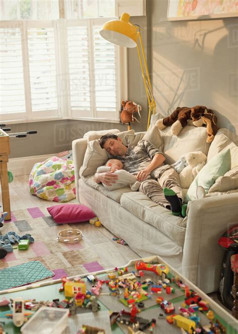 Exhausted Father And Baby Son Sleeping On Sofa In Messy Living Room