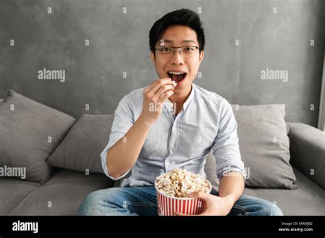 Portrait Of A Happy Young Asian Man Eating Popcorn While Sitting On A