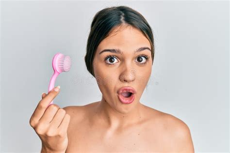 Young Brunette Woman Using Facial Exfoliating Brush Scared And Amazed