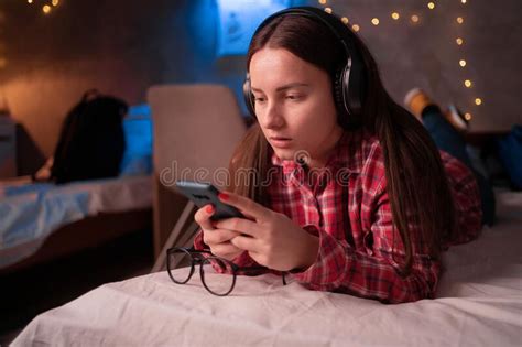 Female College Student Wearing Headphones Lies On Bed In Dormitory With