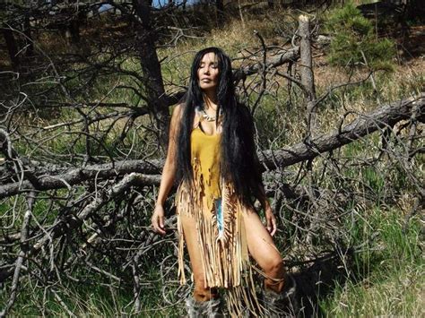 a woman with long black hair wearing a native style dress standing in front of a fallen tree