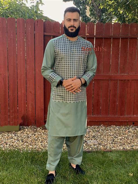 A Man Standing In Front Of A Red Fence Wearing A Green Outfit And Black