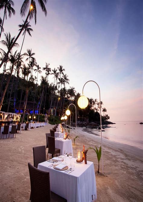 An Outdoor Dining Area On The Beach At Dusk With Palm Trees And Water