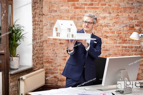 Mature Male Architect Examining Model House While Standing By Desk In