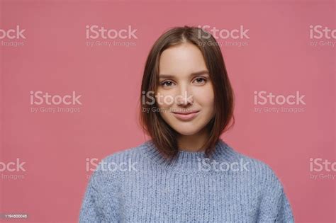 어두운 머리를 가진 아름다운 성인 여성 차분한 얼굴 표정 니트 스웨터를 입고 분홍색 배경에 대해 스튜디오에서 포즈를 취하고 겨울 동안 즐거운 것을 생각하고 부드러운 모습을