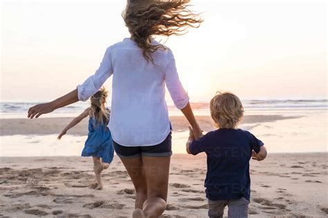 Mother Holding Hand Of Son While Running With Daughter In Background At Beach Stock Photo