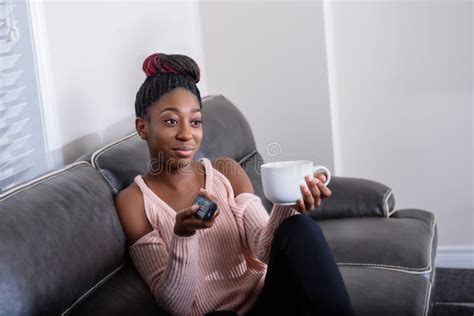 uma mulher afro americano nova que senta se no sofá com a tevê remota e que olha fixamente na