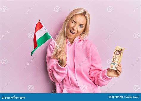 Young Blonde Woman Holding Hungary Flag And Forint Banknotes Winking Looking At The Camera With