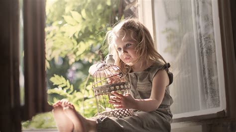 Cute Little Girl Is Sitting Near Window With Bird And Cage On Lap