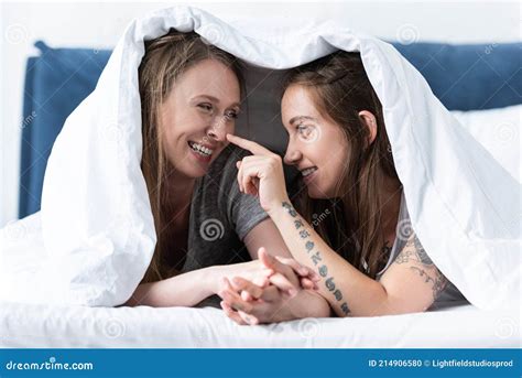 Two Happy Lesbians Holding Hands While Lying Under Blanket On Bed