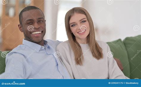 Interracial Couple Sitting On Sofa And Smiling At Camera Stock Image