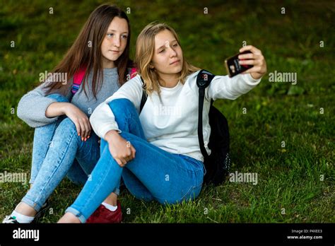 Two Girl Friends Schoolgirl Summer In Park In Nature Sit On The Grass Backpacks In His Hands