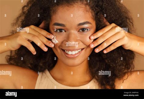 Smiling Dark Skinned Woman With Curly Afro Hair Applies Face Cream On Cheeks With Both Hands