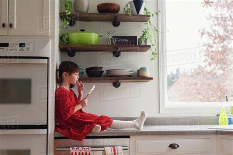 Girl Sitting On Kitchen Counter With A Spatula Eating Frosting Stock