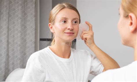 Beautiful Woman Is Getting Massage Face Using Jade Stone In Front Of The Mirror At Home Stock