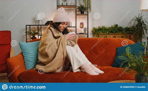 Sick Unhealthy Woman Wear Hat Wrapped In Plaid Sit Shivering From Cold On Sofa Drinking Hot Tea