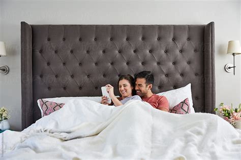 Young Millennial Couple In Bed Cuddling And Texting On A Cell Phone