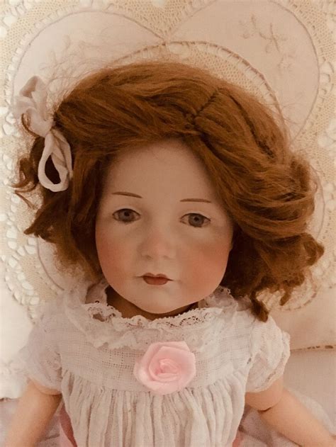 An Old Doll With Red Hair Laying On A White Bed Next To A Lace Doily