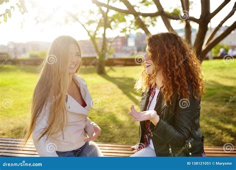 Two Beautiful Young Women Talking While Sitting On A Bench At Sunny